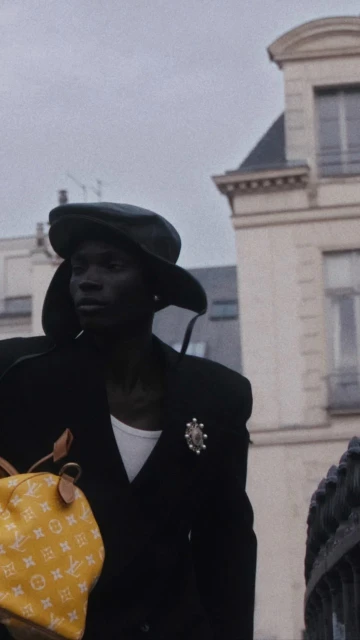 African American man in a black coat and cap walks outside with a yellow Louis Vuitton bag, with a building in the background.