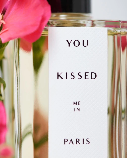 Perfume bottle with a label that reads 'You Kissed Me In Paris' next to pink flowers, capturing a blend of fragrance and floral elegance.