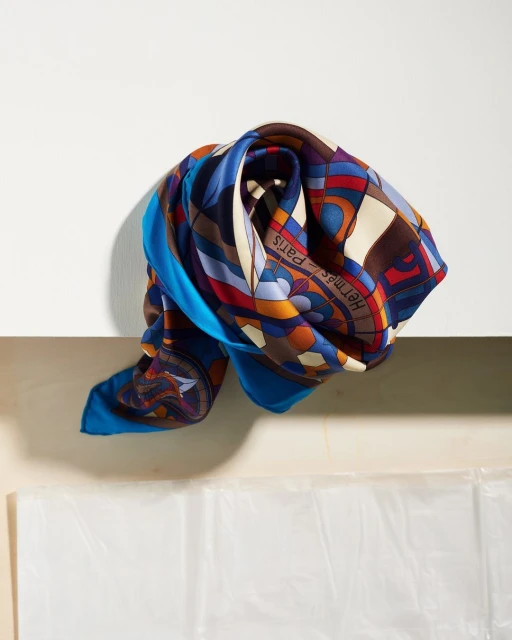 Elegant silk scarf in blue, brown, and red, with geometric patterns and the Hermes Paris logo, draped over a white surface.