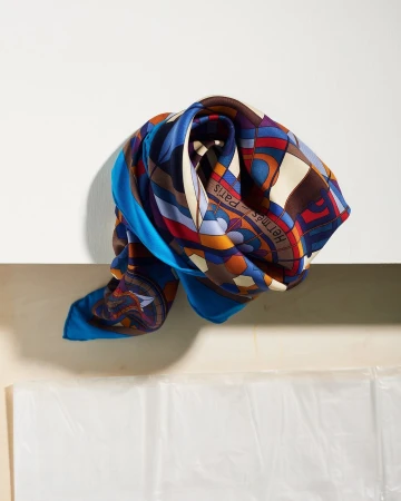 Elegant silk scarf in blue, brown, and red, with geometric patterns and the Hermes Paris logo, draped over a white surface.