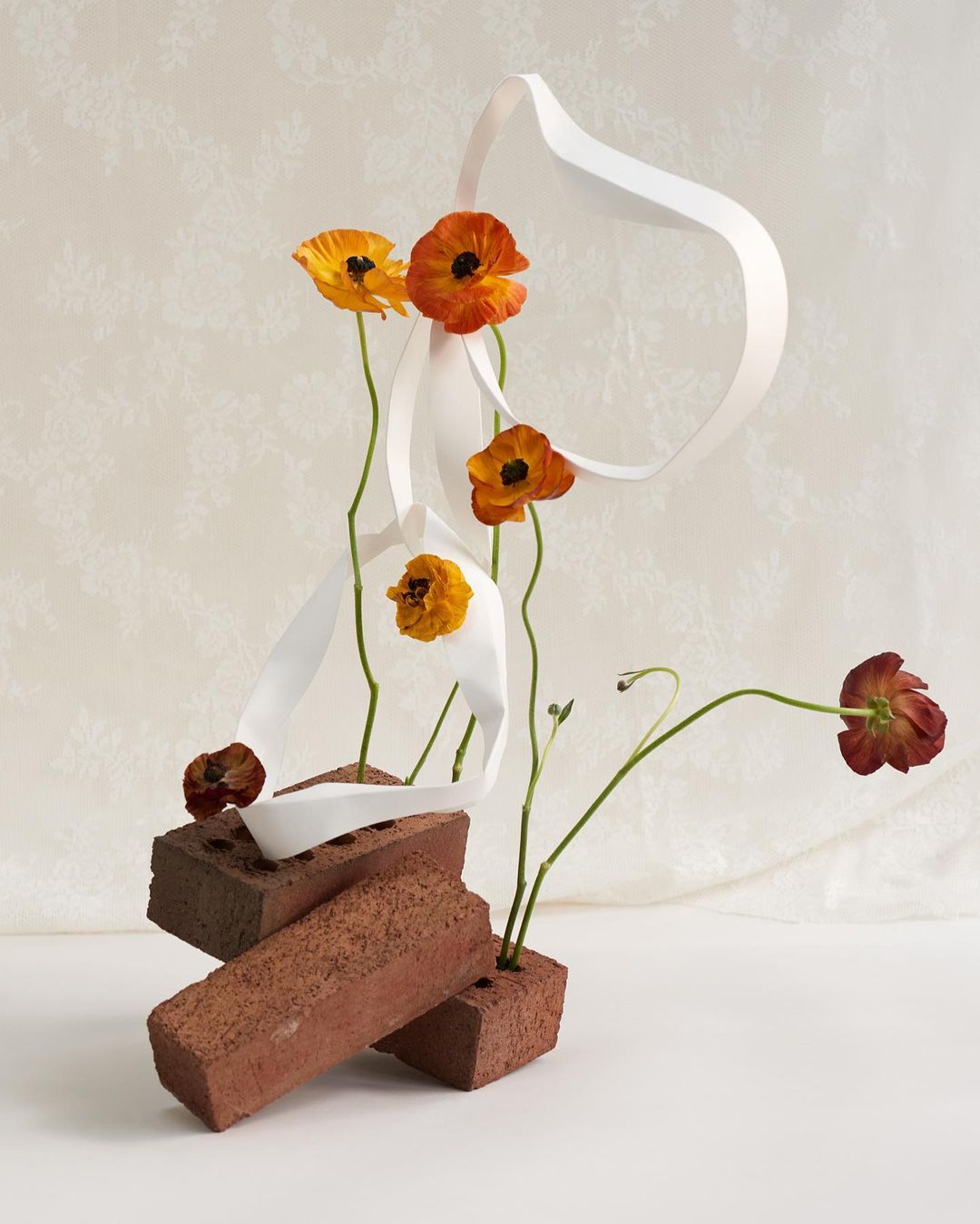 Still life of orange and yellow flowers arranged in red bricks, paired with a flowing white ribbon against a white patterned background.