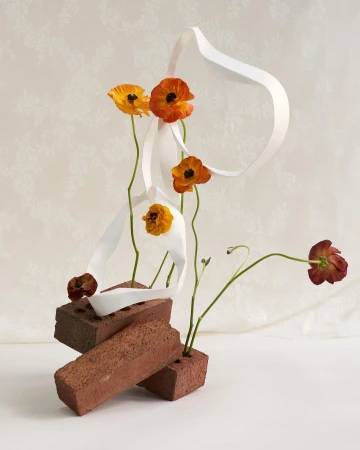 Still life of orange and yellow flowers arranged in red bricks, paired with a flowing white ribbon against a white patterned background.