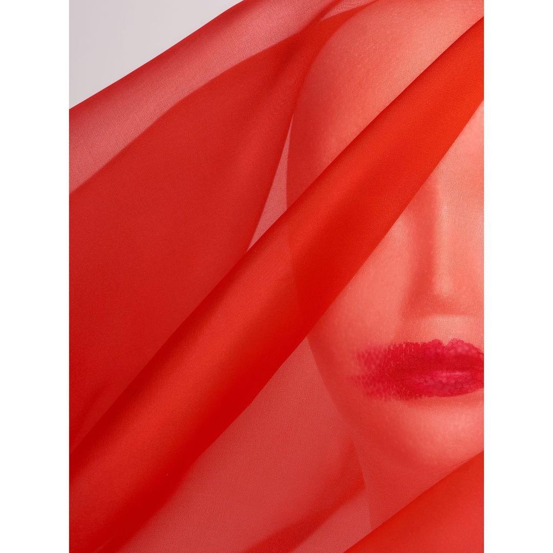 Portrait of woman's face partially obscured by flowing red fabric, lips emphasized with bright red lipstick against a white background.