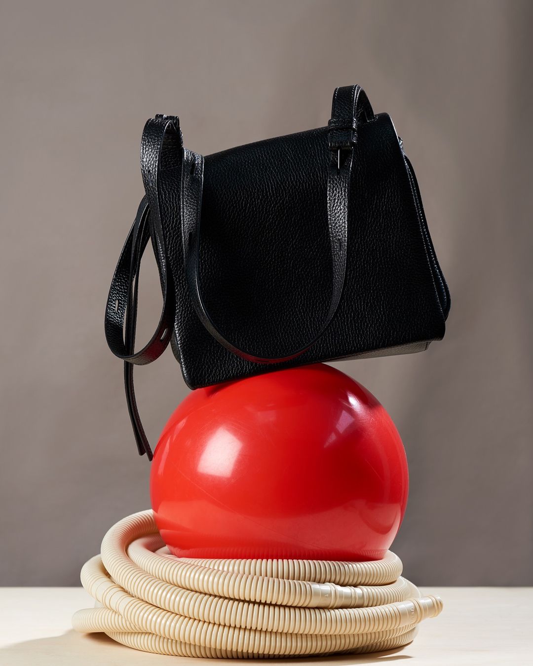 Still life studio shot of a black purse resting atop a red spherical object and beige corrugated tube on a light surface.