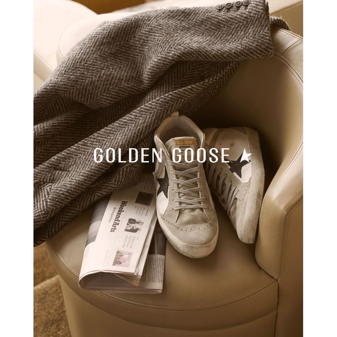 Golden Goose sneakers, newspaper, and grey herringbone jacket on tan leather chair indoors in soft lighting, displaying casual luxury.