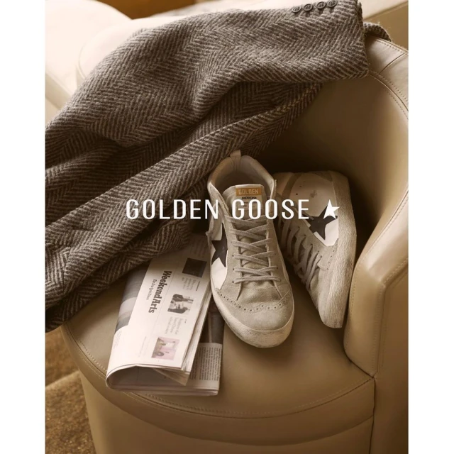 Golden Goose sneakers, newspaper, and grey herringbone jacket on tan leather chair indoors in soft lighting, displaying casual luxury.