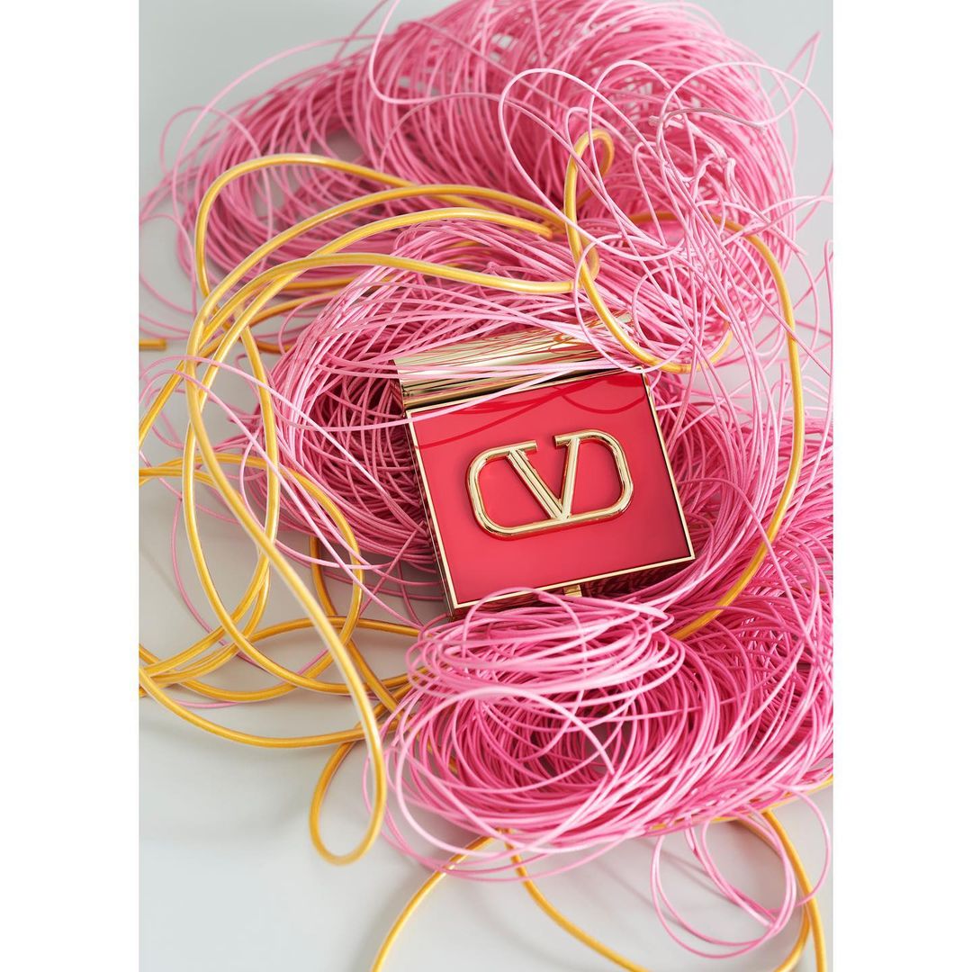 Cosmetic product with a red case and a gold logo surrounded by pink and yellow strings on a white surface, product photography.