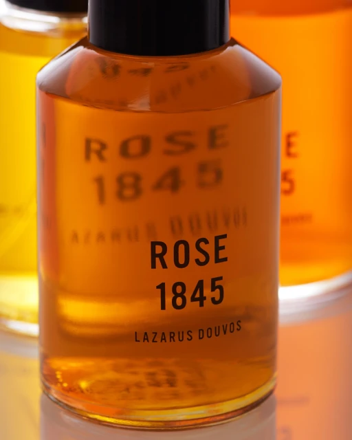 Close-up of Lazarus Douvos ROSE 1845 fragrance bottles filled with amber liquid and dark caps, displayed on a reflective surface.
