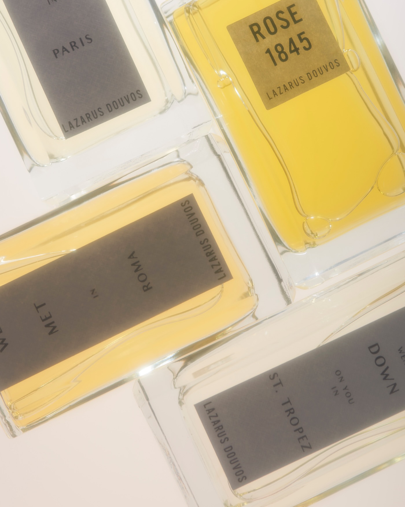 Four perfume bottles with gold labels from Lazarus Douvos, showcasing different scents like Rose 1845, Paris, Roma, and St. Tropez.
