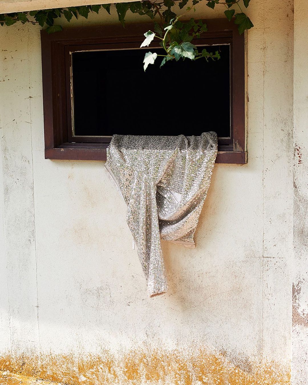 Close-up of a silver sequined gown draped over a dark-framed window in a weathered cream-colored wall, illuminated by soft, natural light.