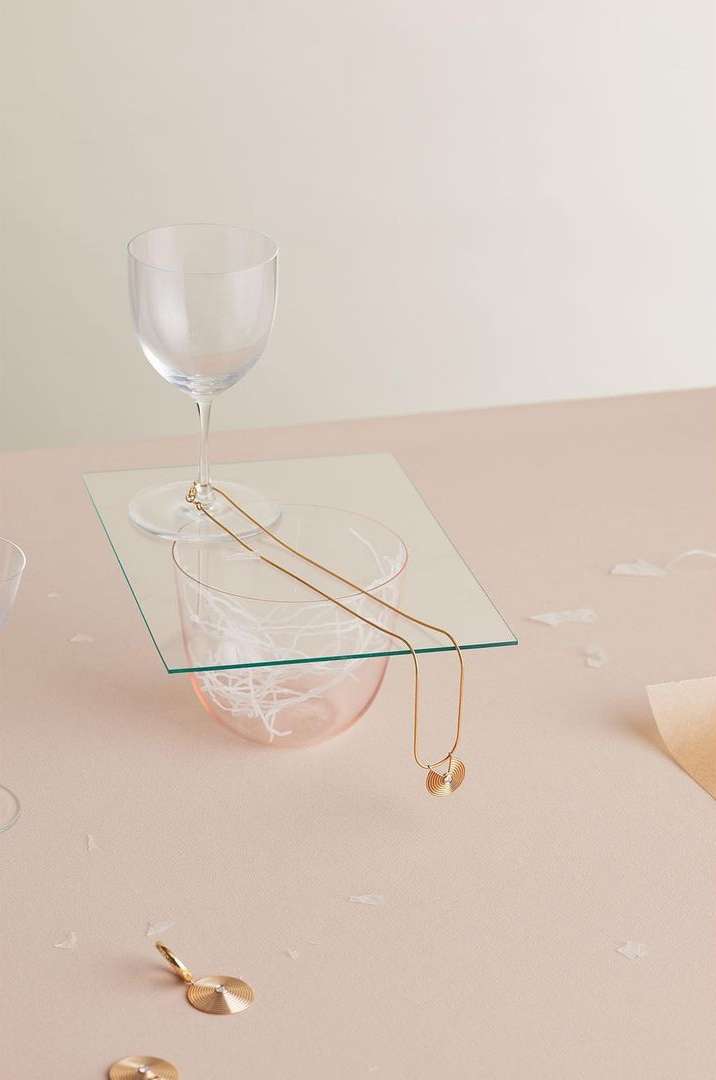 Elegant composition of a gold necklace draped over glassware atop a pink surface, creating a minimalist, high-end jewelry showcase.