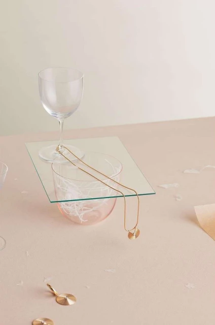 Elegant composition of a gold necklace draped over glassware atop a pink surface, creating a minimalist, high-end jewelry showcase.