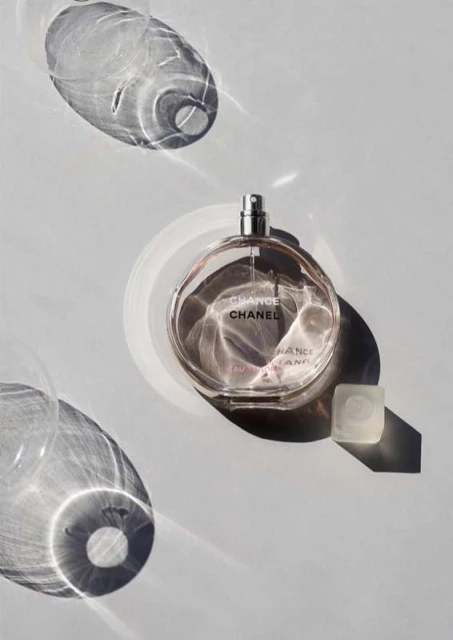 A bottle of Chanel Chance perfume sits on a white surface with geometric shadows, showcasing product photography and elegant simplicity.