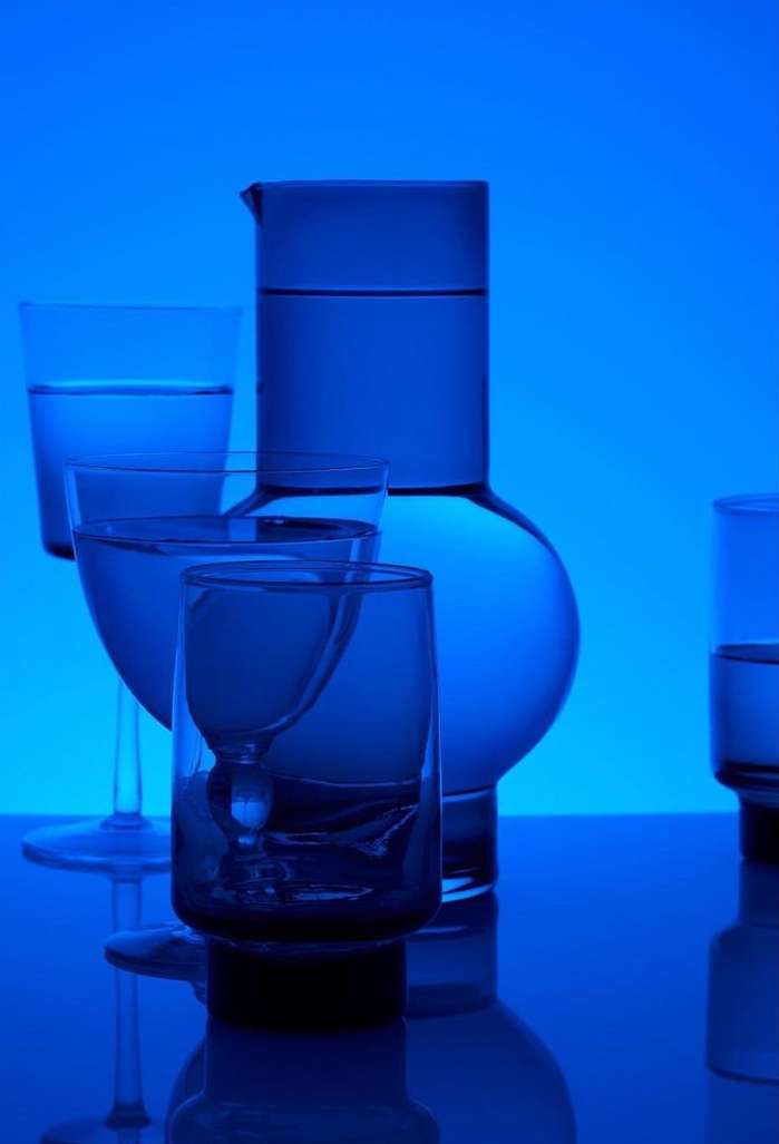 Still life of a clear glass water pitcher and a variety of drinking glasses filled with liquid, set against a vibrant blue background.