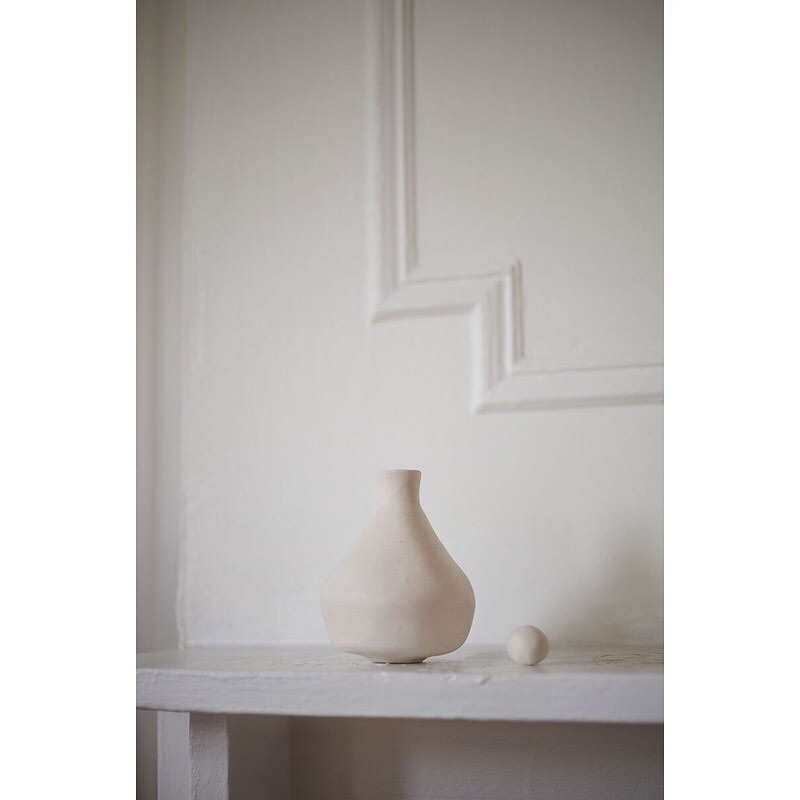 Minimalist interior photo of a light beige ceramic vase and a small sphere sitting on a white shelf against a decorative white wall.