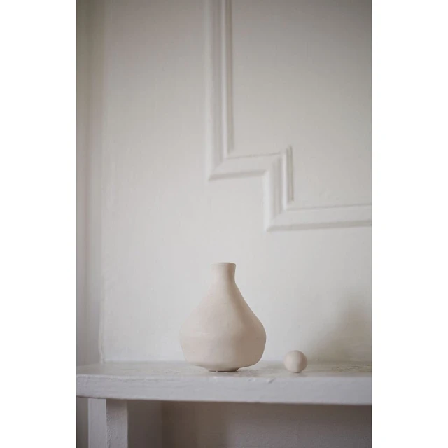Minimalist interior photo of a light beige ceramic vase and a small sphere sitting on a white shelf against a decorative white wall.