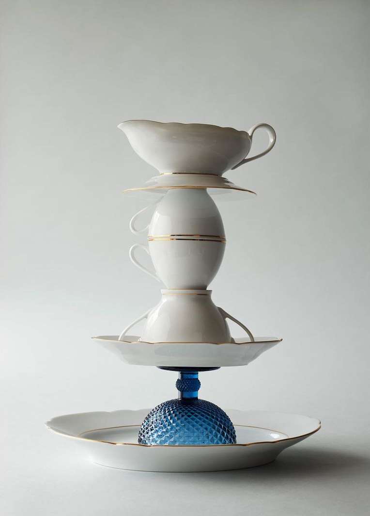 Tower of white fine china teacups and saucers, supported by a blue textured glass base on a white background, studio lit.