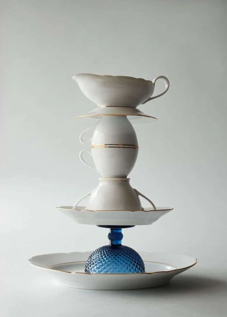 Tower of white fine china teacups and saucers, supported by a blue textured glass base on a white background, studio lit.