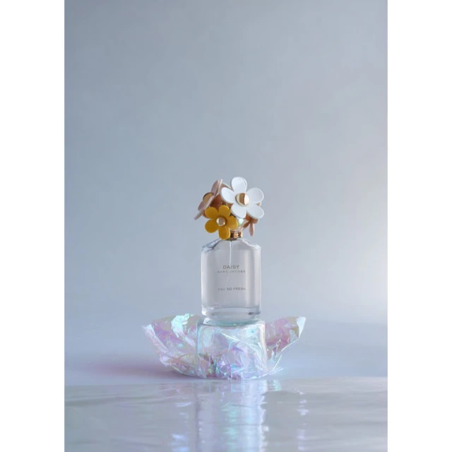 Still life photograph of a bottle of Marc Jacobs Daisy Eau So Fresh perfume topped with daisy decorations, on a pearlescent surface.