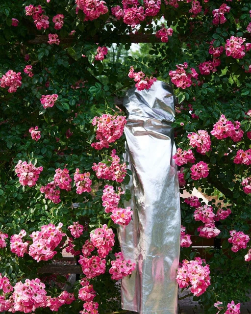 Pink roses cover an archway and a tall object wrapped in shiny silver material, creating a unique floral display in natural light.