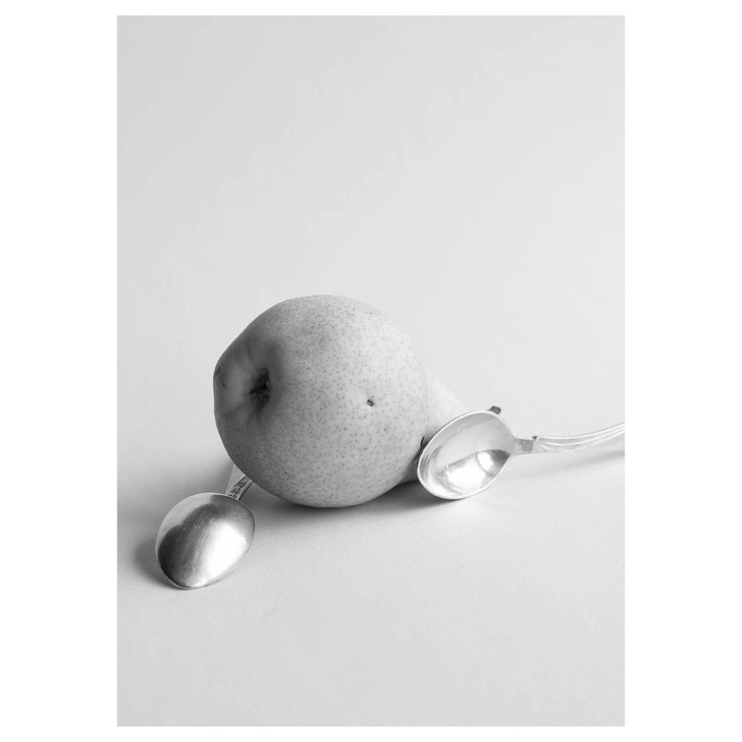 Black and white still life of a pear centered between two silver spoons against a pale background, creating contrast and visual interest.
