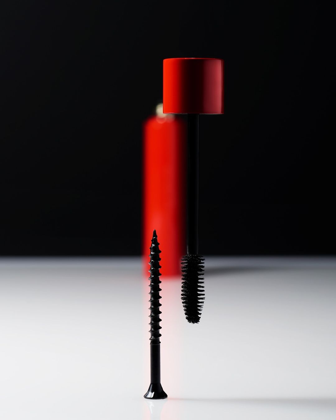Still life studio shot of a black screw standing next to a black mascara brush and red makeup bottle, on a white surface against black.