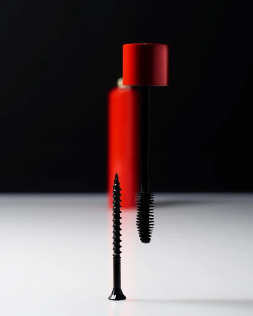 Still life studio shot of a black screw standing next to a black mascara brush and red makeup bottle, on a white surface against black.