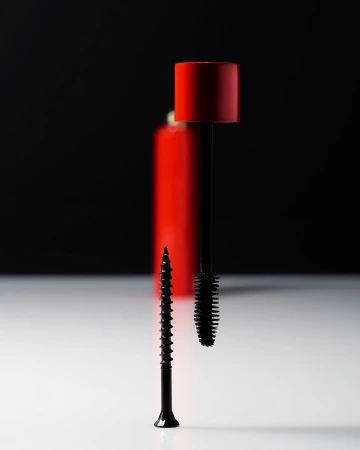 Still life studio shot of a black screw standing next to a black mascara brush and red makeup bottle, on a white surface against black.