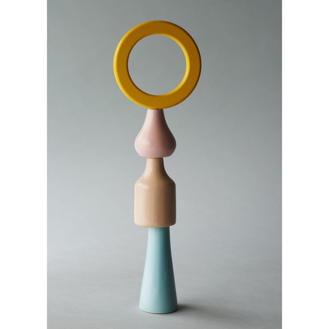 Abstract sculpture featuring a blue cone, beige cylinder, pink drop shape, and yellow ring against a plain grey background.