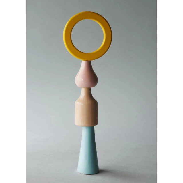 Abstract sculpture featuring a blue cone, beige cylinder, pink drop shape, and yellow ring against a plain grey background.