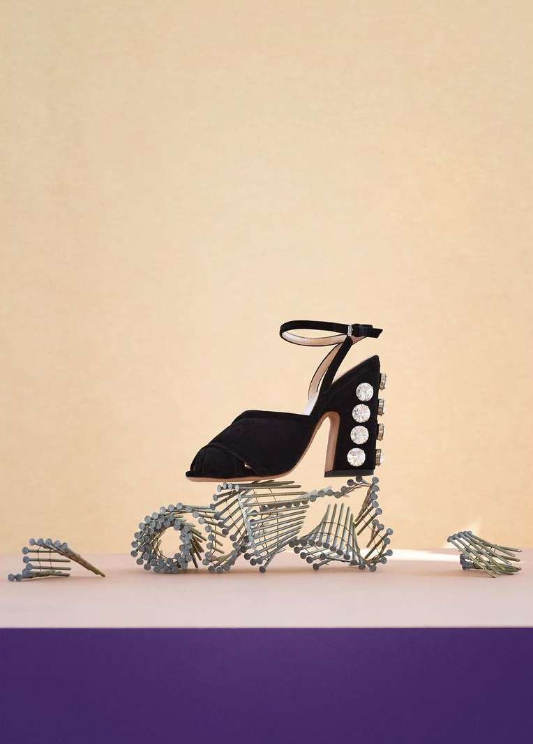 Black high-heeled sandal with rhinestone details on a purple surface, balanced atop a sculpture of metal screws against a beige backdrop.