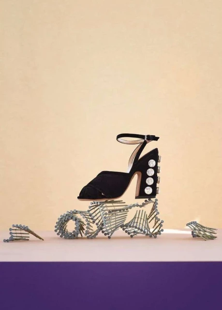 Black high-heeled sandal with rhinestone details on a purple surface, balanced atop a sculpture of metal screws against a beige backdrop.