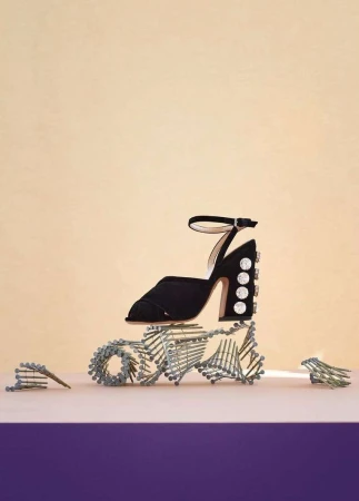 Black high-heeled sandal with rhinestone details on a purple surface, balanced atop a sculpture of metal screws against a beige backdrop.
