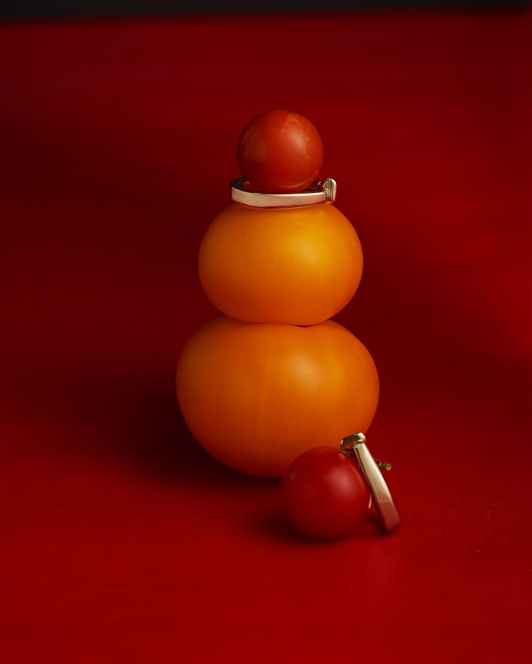 Three tomatoes are stacked on top of each other, adorned with silver and gold rings, against a vibrant red backdrop.