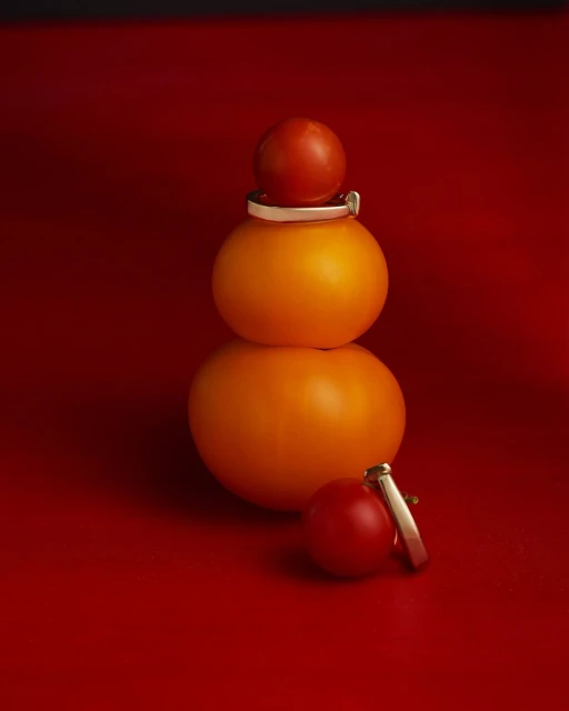 Three tomatoes are stacked on top of each other, adorned with silver and gold rings, against a vibrant red backdrop.