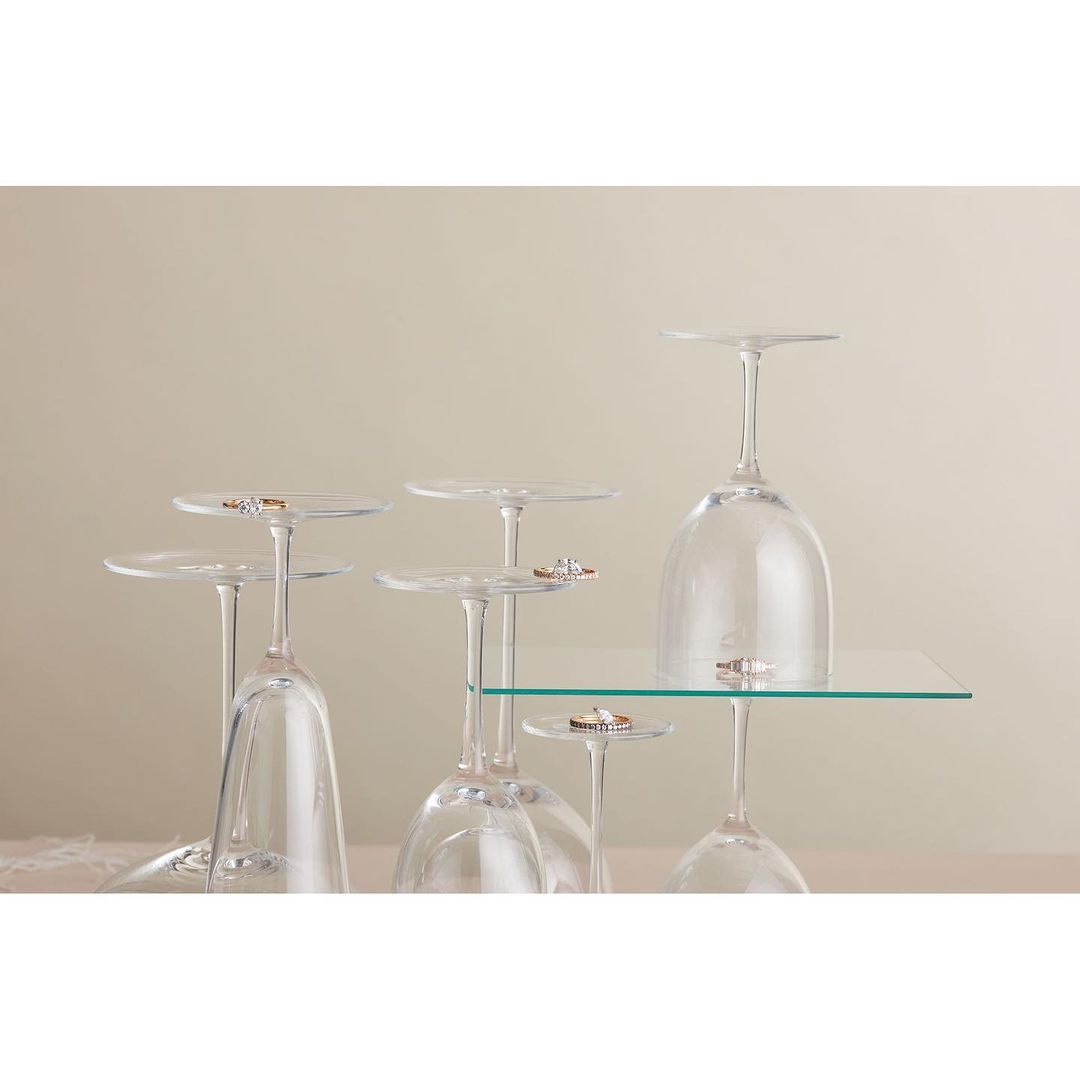 Elevated view of six upside-down wine glasses holding wedding bands, set against a neutral background, some glasses on a clear glass shelf.