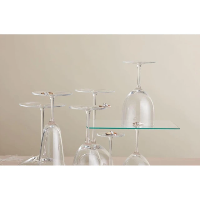 Elevated view of six upside-down wine glasses holding wedding bands, set against a neutral background, some glasses on a clear glass shelf.