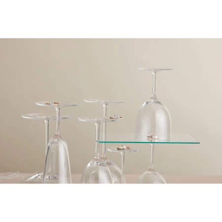 Elevated view of six upside-down wine glasses holding wedding bands, set against a neutral background, some glasses on a clear glass shelf.