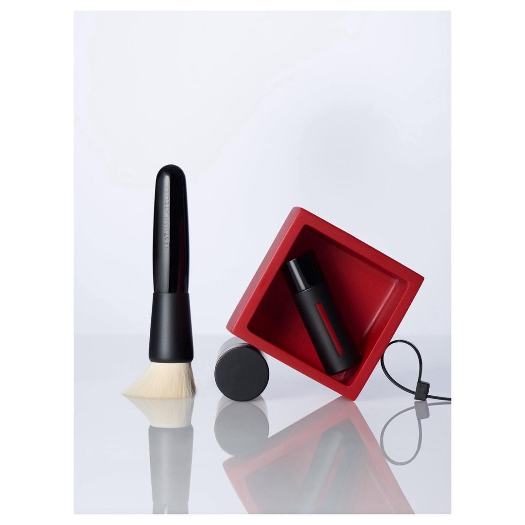 Cosmetic product shot: makeup brush, black bottle, and red container on a white reflective surface, studio setting, minimalist.