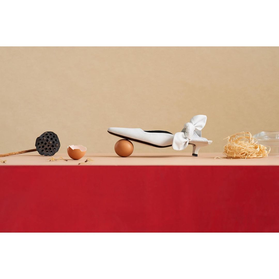 White heeled shoe with a bow on a brown egg with a neutral background next to a lotus pod and noodles.