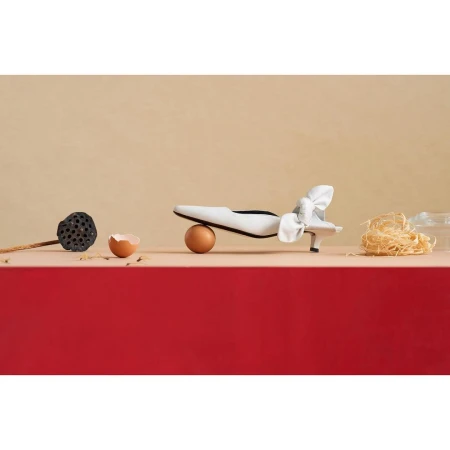 White heeled shoe with a bow on a brown egg with a neutral background next to a lotus pod and noodles.
