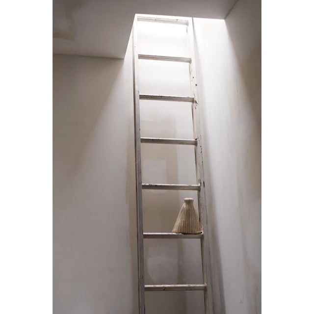 A tall, white wooden ladder extends upward, with a woven lampshade resting on one of its rungs, set against a plain white wall.