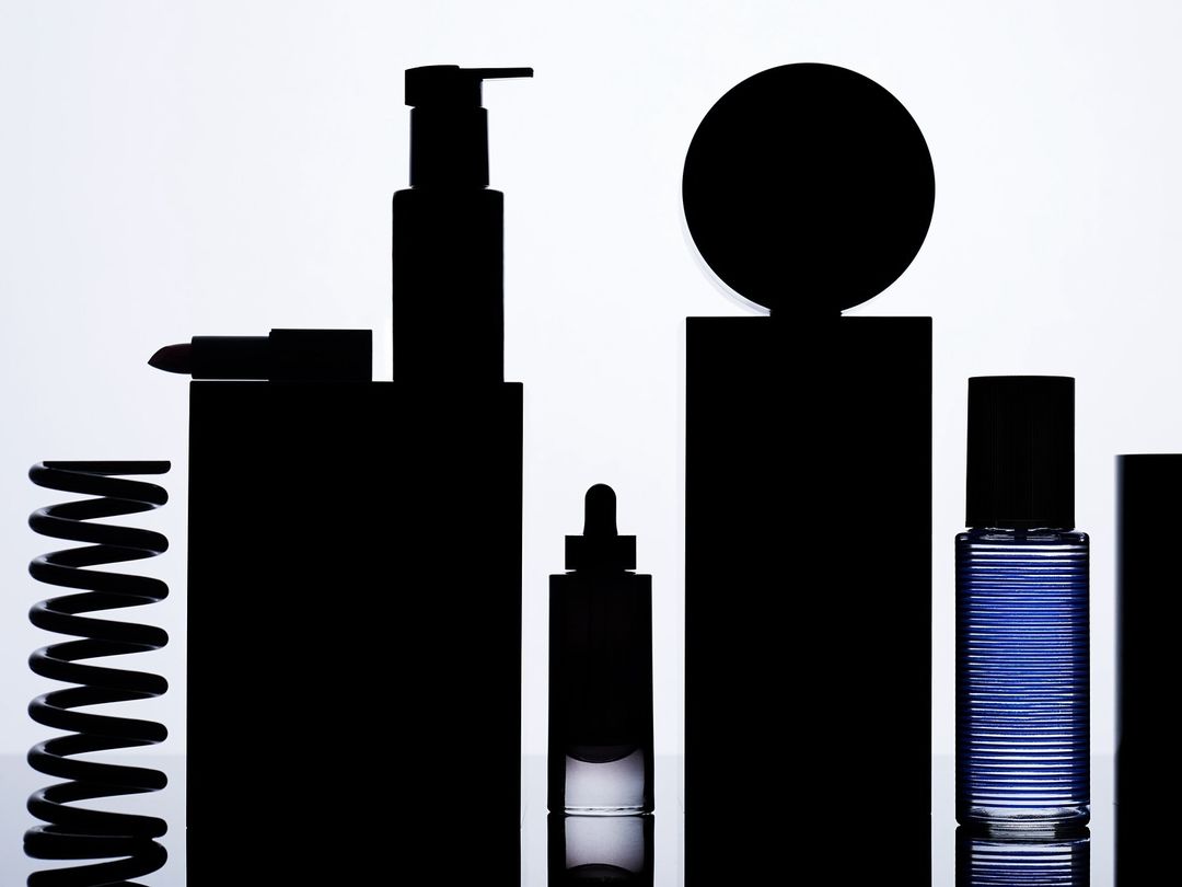 Silhouette of assorted cosmetic containers and a spring on a reflective surface against a graduated white backdrop, studio lighting.