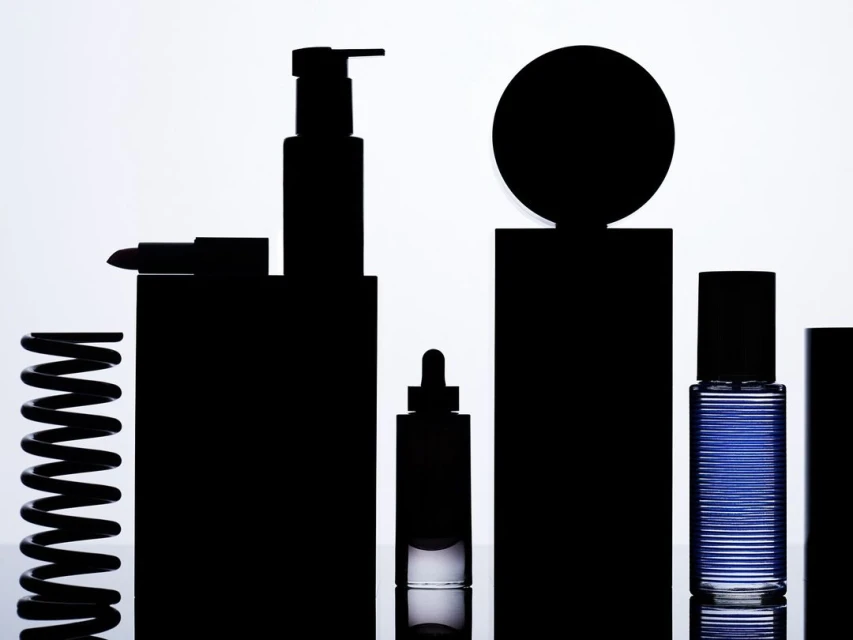 Silhouette of assorted cosmetic containers and a spring on a reflective surface against a graduated white backdrop, studio lighting.