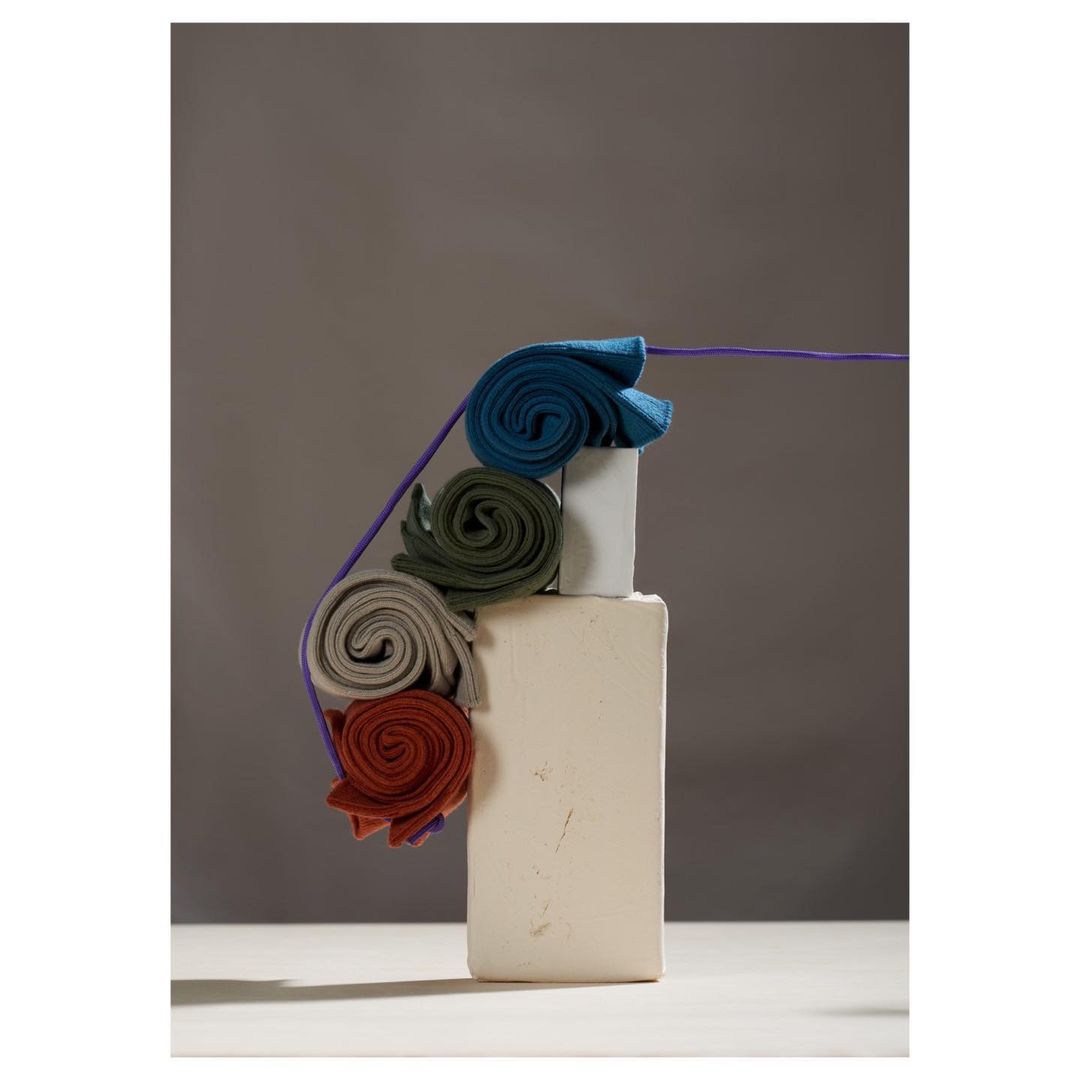Stack of rolled colorful fabrics balanced on white blocks, studio shot with a purple cord, neutral background, minimalist.
