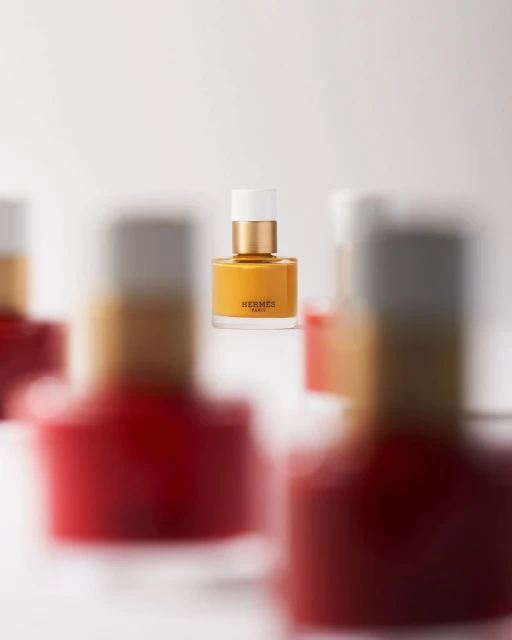 Still life shot of a yellow Hermès Paris nail polish bottle in focus, surrounded by several blurred red and silver nail polish bottles.