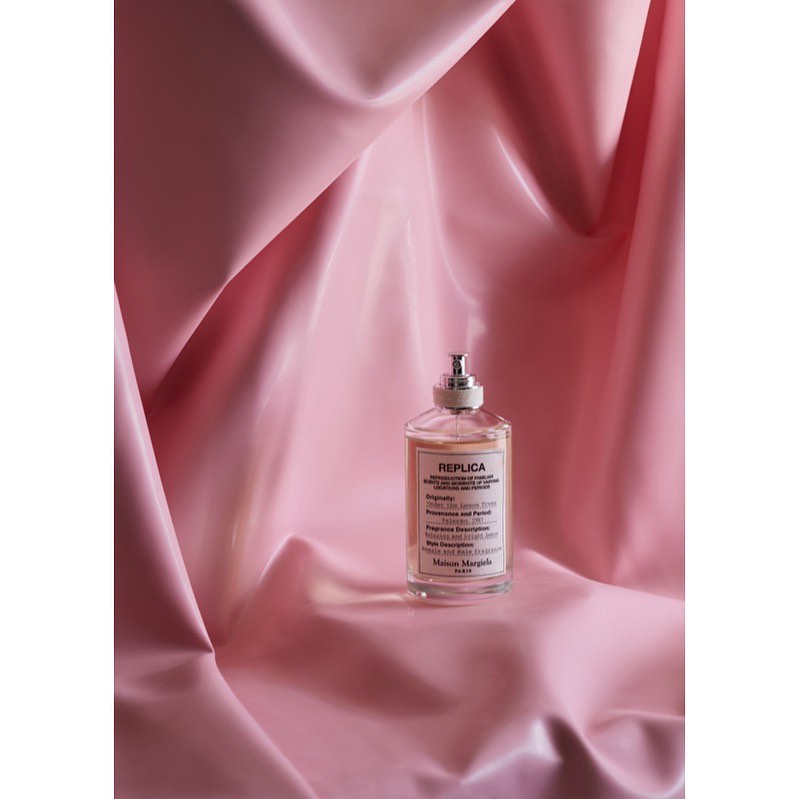 Still life studio shot of REPLICA perfume bottle with a silver cap, label, and light pink liquid, displayed on a draped light pink fabric.