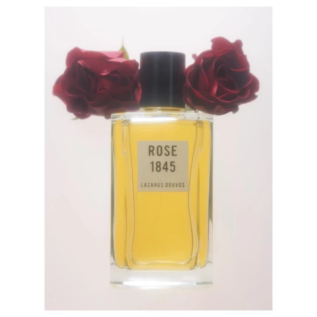 An artful product shot features a bottle of ROSE 1845 Lazarus Douvos perfume, flanked by two dark red roses, set against a soft white background.