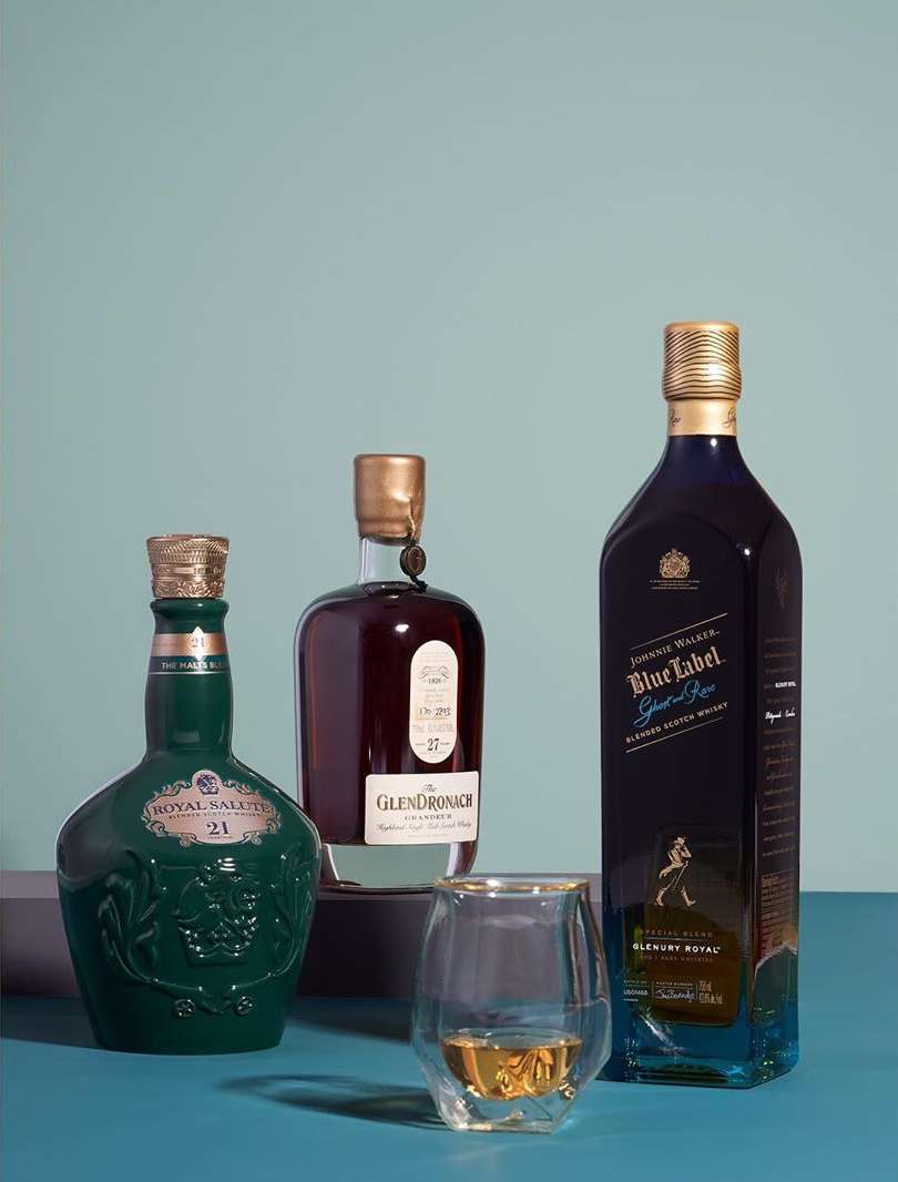 Still life shot of three whisky bottles (Royal Salute, Glendronach, Johnnie Walker Blue Label) and a glass of whisky on a blue background.