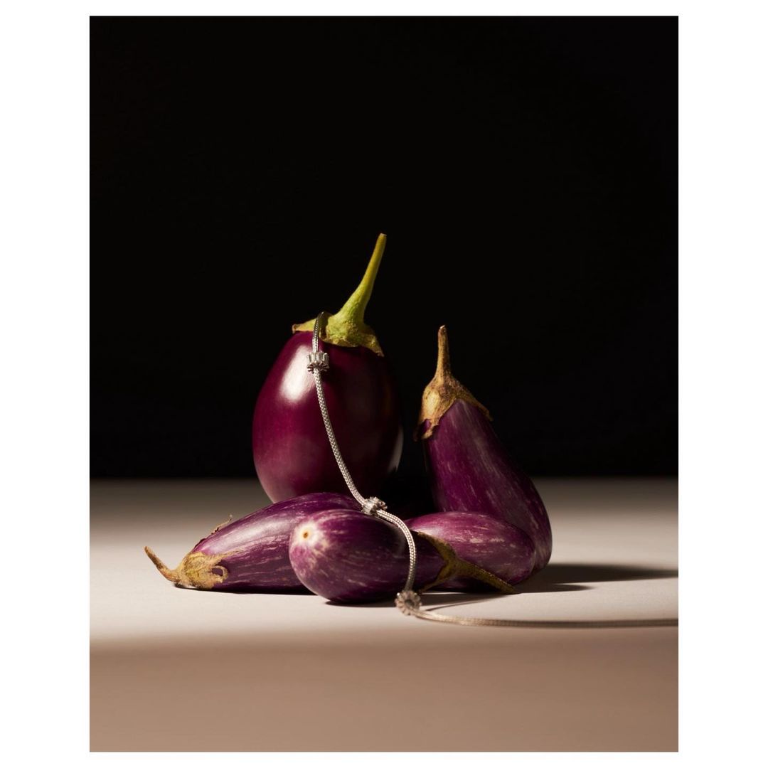 Studio shot of several eggplants on a table, with a silver chain draped across the vegetables against a dark brown background.